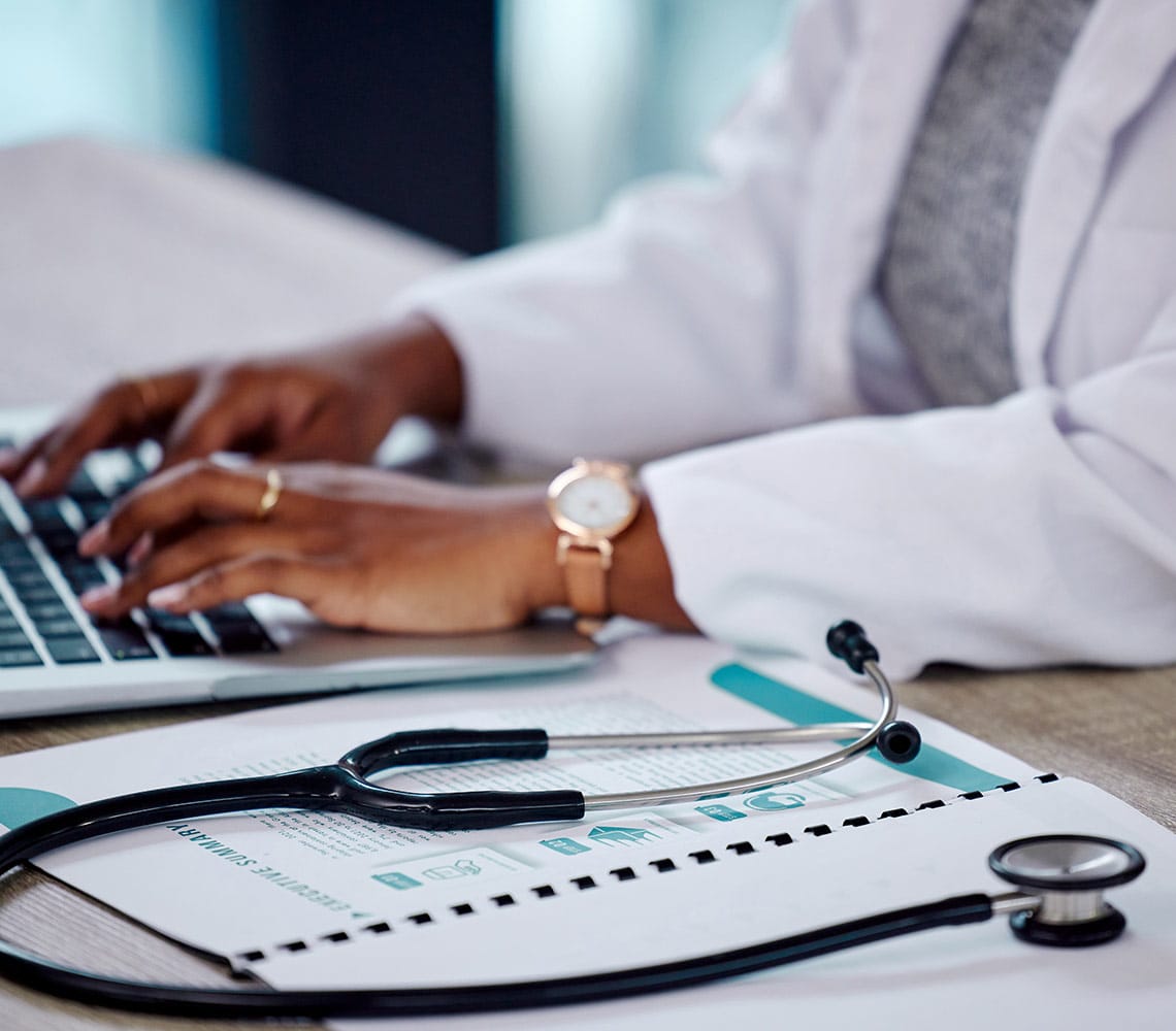 A physician working on a laptop while a stethoscope rests on the table.